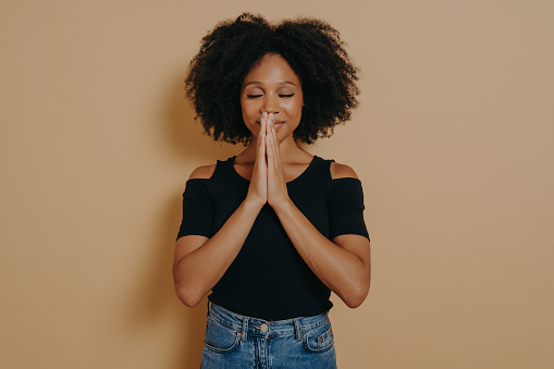 Afro American young woman praying and holding hands in prayer gesture standing isolated on dark beige studio background with copy space, smiling with eyes closed, Praying and hope concept