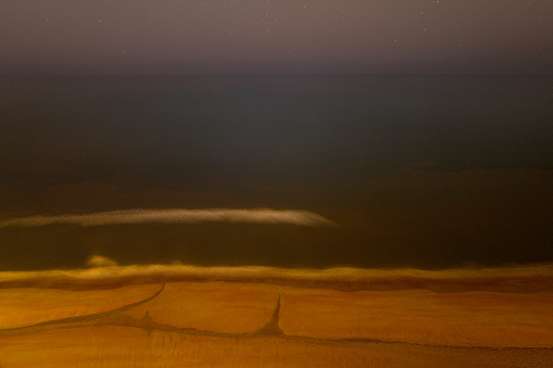 Foggy night ocean and beach seascape, long exposure aerial photography, earth-toned golden-color seashore, white lines of foamy sea waves, dimly illuminated sky with stars