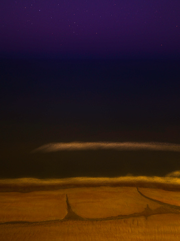 Night ocean and beach seascape, long exposure aerial photography, earth-toned golden-color sand, white lines of foamy sea waves, purple sky with stars