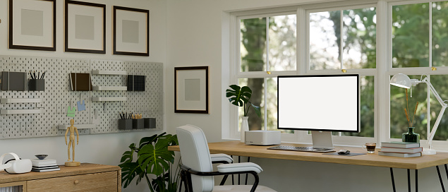 Beautiful white home office interior design with computer mockup on woods desk against the window with nature view, armchair, pegboard and blank frame mockup on white wall. 3d render, 3d illustration