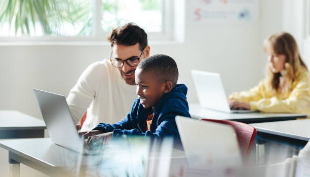 Technology education: Teacher assisting student with computer science lesson Male teacher helps a young boy with computer-based learning in a classroom setting. Child tutor providing a lesson in an elementary school, with a focus on coding and basic digital literacy. education stage stock pictures, royalty-free photos & images