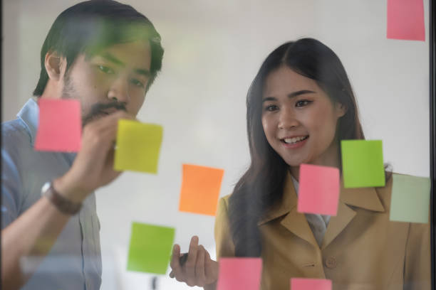 istock Casual creative happy two smiling Asian business people plan strategy analysis and brainstorm by putting post-it sticker notes to share an idea for a startup project on a glass board. 1468458918