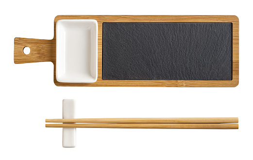 Bamboo sushi serving set cutout. Wooden board with black slate tray, porcelain soy sauce dish and pair of chopsticks on a rest isolated on a white background. East Asian tableware. Top view.