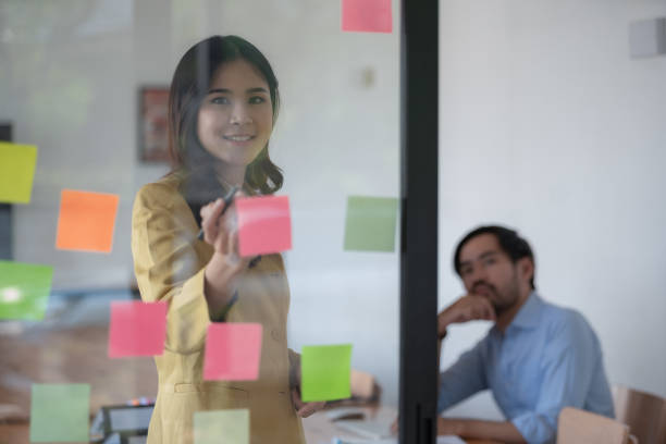 istock Casual creative happy two smiling Asian business people plan strategy analysis and brainstorm by putting post-it sticker notes to share an idea for a startup project on a glass board. 1468461020