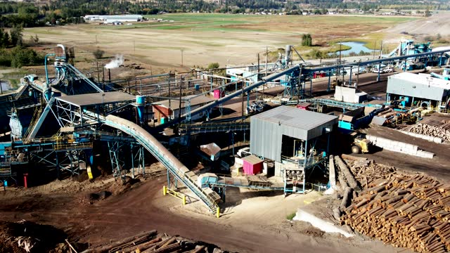dolly forward zoom drone shot of a blue Sawmill Processing Plant with log piles in the background in a desert environment with heavy equipment moving