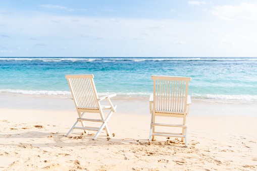 Two white chairs on the beach. Blue ocean and clear sky with few clouds on the background. Shot taken on Bali, Bukit.
