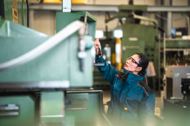 Asian Female Factory Worker Operating An Industrial Machine Mid adult female blue collar worker operating an industrial machine in a factory. She is wearing protective clothes and eyeglasses. Pressing buttons on a machine. machine stock pictures, royalty-free photos & images