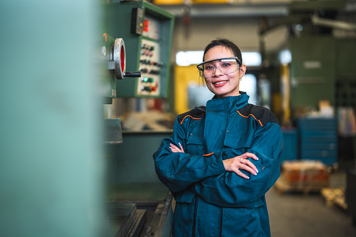 Portrait of a mid adult female blue collar worker industrial machine operator in a factory with arms crossed. She is wearing protective clothes and eyeglasses.