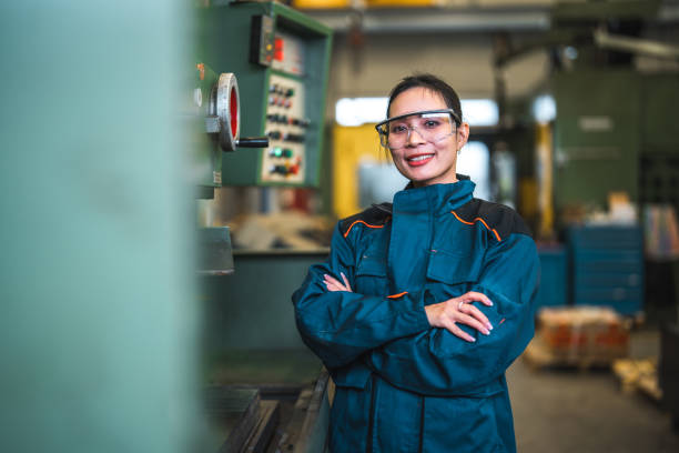 Asian Female Blue Collar Worker With Arms Crossed At Work Portrait of a mid adult female blue collar worker industrial machine operator in a factory with arms crossed. She is wearing protective clothes and eyeglasses. asian female engineer stock pictures, royalty-free photos & images
