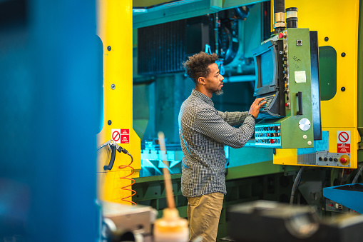 Mid adult Black blue collar worker operating an industrial CNC machine in a factory. He is wearing casual clothes and pressing buttons on a machine and programming it.