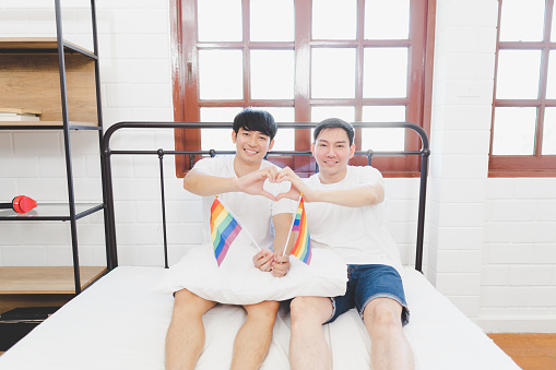 Gay couple waving lgbt rainbow flag and Romantic Hand made heart-shaped hand to make love happily. indoors on bed at home. Attractive romantic male lgbt couple sit on bed in bedroom in morning.