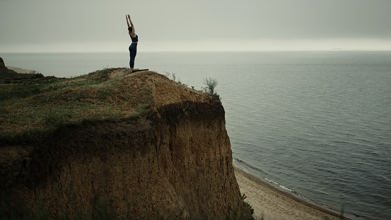 Sporty young woman making yoga exercise standing on top sandy hill. Slim sportswoman stretching on nature wearing dark sportswear. Flexible girl doing gymnastic outdoors cloudy day. Sport concept.