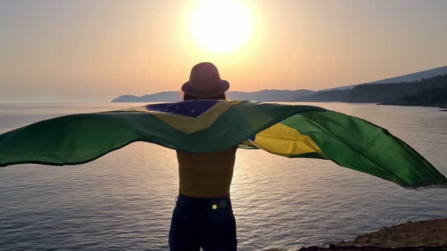 Young Caucasian woman waving a Brazilian flag on the seaside during sunset