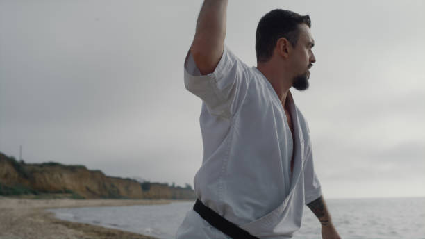 Bearded kungfu man practicing martial arts on sandy beach cloudy morning close up. Sporty athlete training fighting exercises overcast weather outdoor. Young karate fighter workout wearing kimono.