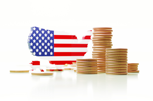 Geographical border of the USA textured with American flag sitting next to coin stacks on white background. Horizontal composition. GDP concept.