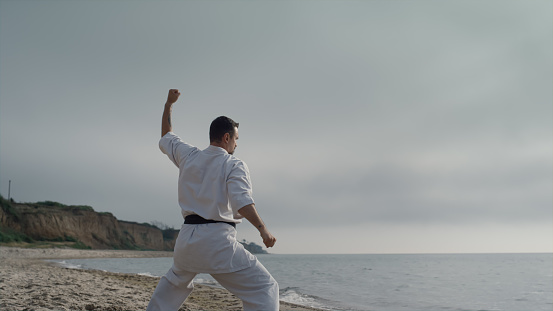 Karate fighter training strength on sandy beach sunny morning. Athletic man standing in fighting position practicing martial arts outdoors. Young sportsman making combat exercises on nature.