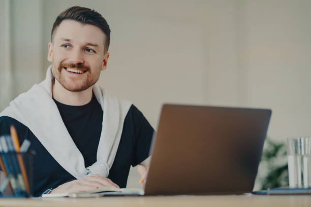 istock Happy bearded man office worker writes organisation plan works remotely makes notes of info sits in front of opened laptop looks away dressed in casual t shirt and jumper tied over shoulders 1468482279