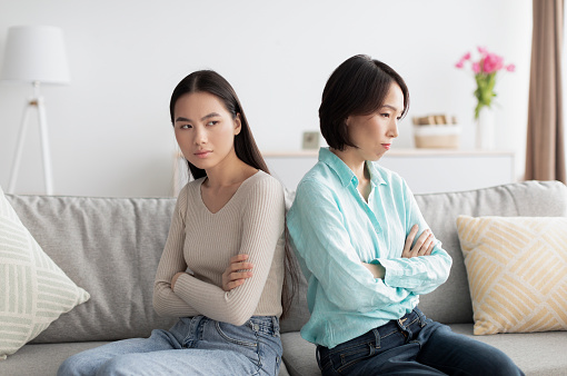 Generations conflict, family problems concept. Offended young Asian woman and her mature mother sitting back to back on couch after argument, not speaking, ignoring each other at home
