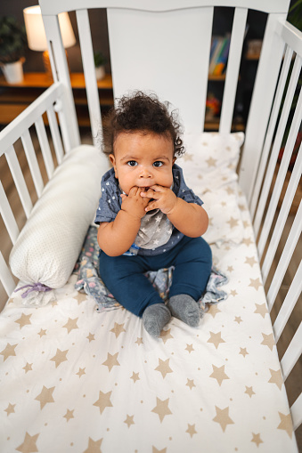 Portrait of multiracial baby while sitting in nursery bed putting hands in mouth