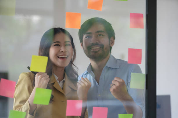 istock Casual creative happy two smiling Asian business people plan strategy analysis and brainstorm by putting post-it sticker notes to share an idea for a startup project on a glass board. 1468487488