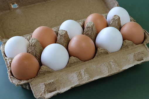 Stock photo showing close-up view of batch of ten mixed white and brown coloured eggs in open disposable cardboard egg box on green background.