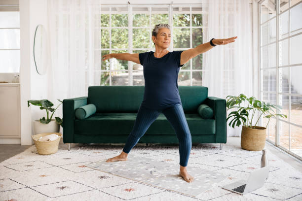 Warrior pose for seniors: Mature woman shows her strength and flexibility as she practices this yoga pose at home Mature woman showing her strength and flexibility as she practices the powerful warrior pose at home. Active senior woman doing yoga in an online fitness class, following a healthy workout routine. warrior position stock pictures, royalty-free photos & images