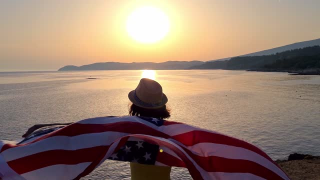 Young Caucasian woman waving a USA flag on the seaside during sunset