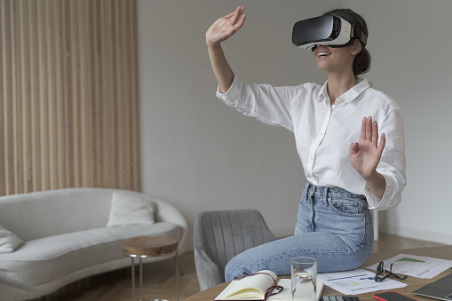 Joyful young business woman sitting on top of desk at home wearing VR glasses, raising hands up while experiencing virtual reality using 3d headset. Technology, innovation and cyberspace concept