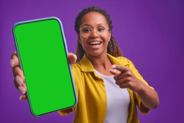 une jeune femme afro-américaine heureuse fait la démonstration d’un téléphone avec un écran vert - isolated on black photos photos et images de collection