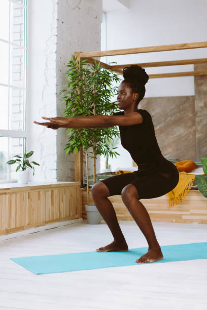 istock Athletic afro american woman squatting with arms extended forward side view, training on gymnastic mat in decorated studio. Sports course, exercise technique, fitness, keeping body fit 1468492994