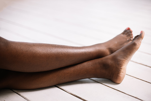 Bare legs of afro american woman closeup, white floor background free copy space. Black person sitting with outstretched legs. Hairy and unshaven legs, new normal and fashion, body positive.