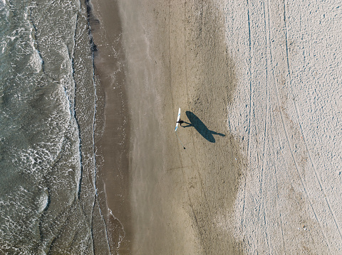 Aerial view of a man walking at the beach with his surfboard. His shadow is visible on the sand.