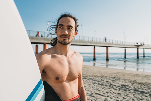 Portrait of a man holding his surfboard on the beach. He's standing on the beach near a pier.