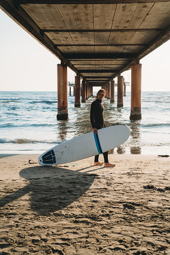 A man is standing on the beach under a pier with his surfboard. He's wearing a wetsuit.