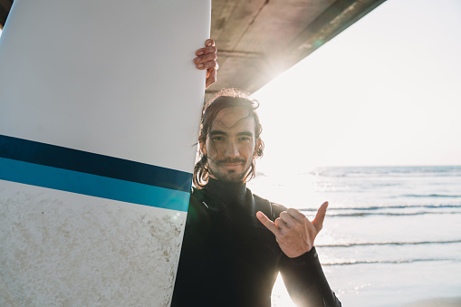 Portrait of a surfer man making the shaka sign to the camera. He's under a pier on the beach.