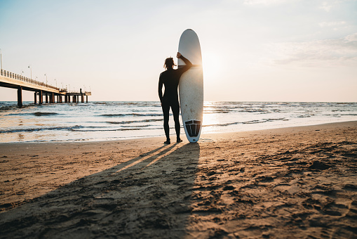 A man is standing on the beach with his surfboard, admiring the sunset