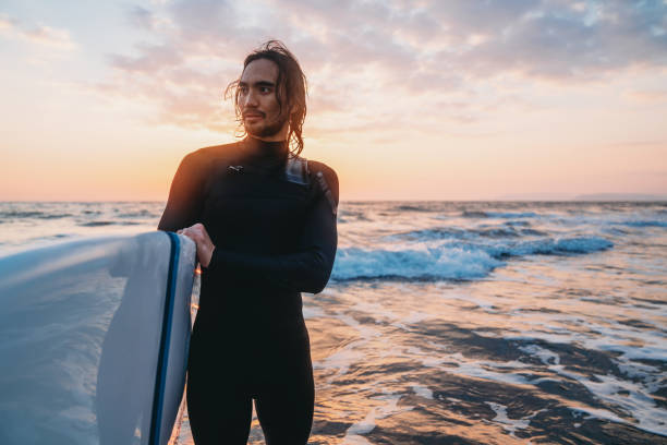 A surfer with his surfboard in the sea at sunset. He's wearing a wetsuit.