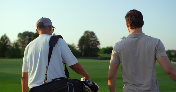 Dad son going fairway course. Two golfers take sport equipment clubs outdoors back view. Unknown golfing players walking together countryside club territory. Family bonding recreation activity concept