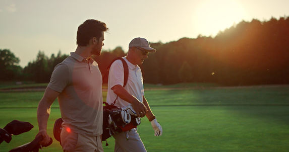 Businessmen walking golf course outside. Two professional players carry clubs in sportswear. Golfing team friends discuss wealthy lifestyle hobby on summer sunset field. Sport partnership concept.