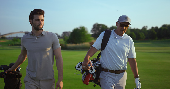 Handsome golf players talk sport hobby. Active men carry golfing putters clubs outdoors. Successful professional golfers walking on green grass club field on summer weekend. Sportsmen together concept