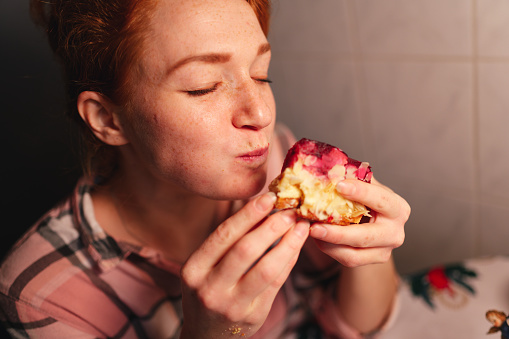 Portrait of a red-haired woman in her kitchen. She treats herself to a piece of raspberry cake. She eats the cake with her hands and has fun.