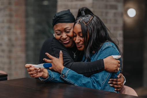 A supportive mature adult black woman who has cancer smiles and embraces her young adult daughter who is holding a pregnancy test. The mother and adult daughter are at home and are happy a baby will be joining their family.
