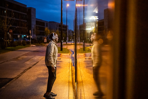 Young man wearing headphones listening to music while standing at bus stop.