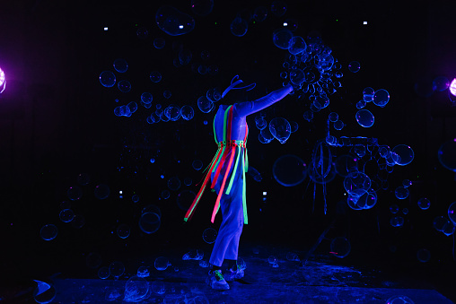 Soap bubble show. The woman artist blows many bubbles at the night party. Female magician makes big bubbles indoors. Actress in stage neon costume. Concept of performance. Neon glowing flying balls.