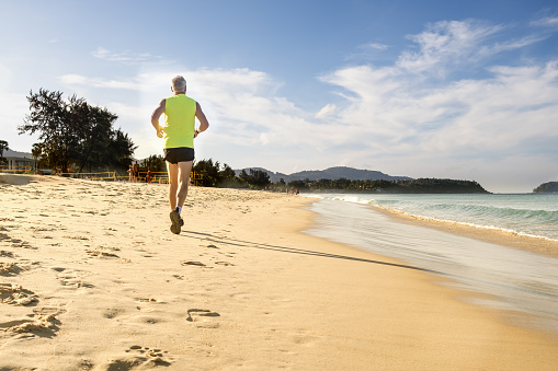 Gray-haired elderly man of athletic build runs along the edge of the sea on a sandy beach. Jogging on the coast. Maintaining physical fitness, playing sports.