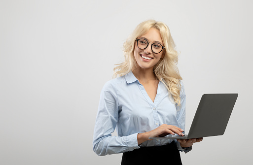 Happy young businesswoman using laptop computer against light grey studio background, looking aside at copy space. Female entrepreneur working with portable pc