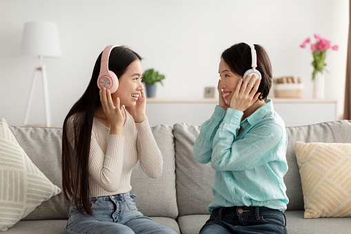 Happy young Asian woman and her mature mother in headphones listening to music together, sitting on sofa at home. Cheerful family enjoying favorite songs together, having fun indoors