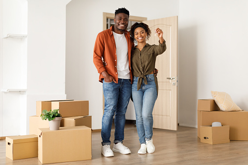 Real Estate Ownership. Joyful Black Couple Showing Key Standing In New House After Relocation, Smiling To Camera Posing Among Moving Boxes. Housing For Young Family