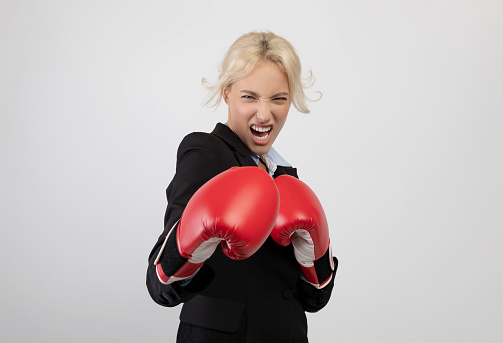 Angry young businesswoman in boxing gloves ready for fight over light grey studio background. Female office worker in formalwear preparing for combat, challenging competitors