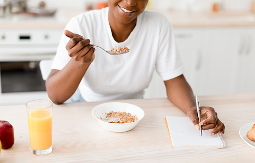 Smiling millennial black female eating porridge at table with cookies, juice and apple, make notes in scandinavian kitchen interior. Health care, proper nutrition at home, breakfast and good morning